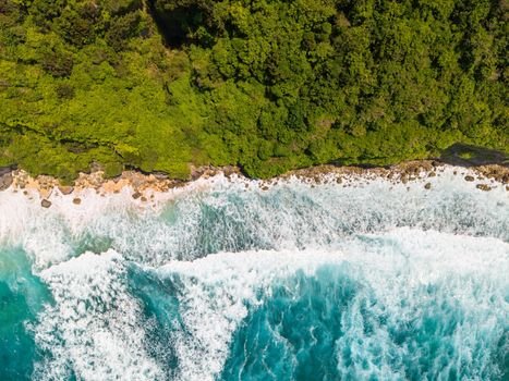 Aerial shot of Bali's coastline with turquoise waves crashing against lush green jungle cliffs.